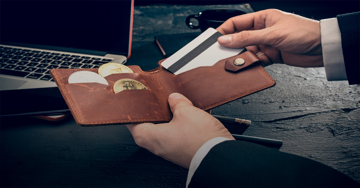Person in a suit holding a leather wallet with physical Bitcoin tokens and a credit card near a laptop on a dark desk