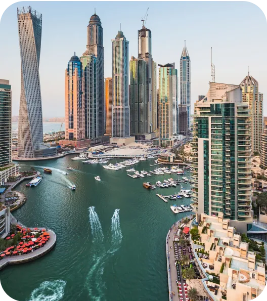 Modern skyscrapers by a marina with boats and yachts in the water, seen from above with city buildings and waterfront paths.