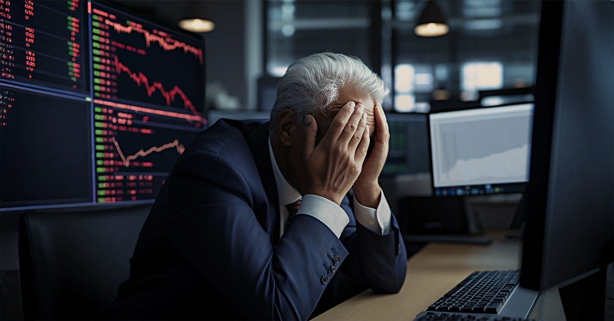 Stressed trader in suit covers face at desk with plummeting stock charts on monitors; symbolizes financial loss and emotional toll of trading.