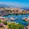 cyprus A scenic harbor with small boats and yachts, surrounded by old-style buildings and distant mountains under a clear blue sky.